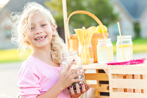 smiling young blonde girl missing teeth next to her lemonade stand