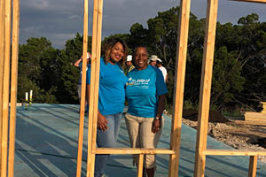 Two women in blue t-shirts stand inside a wooden house frame.