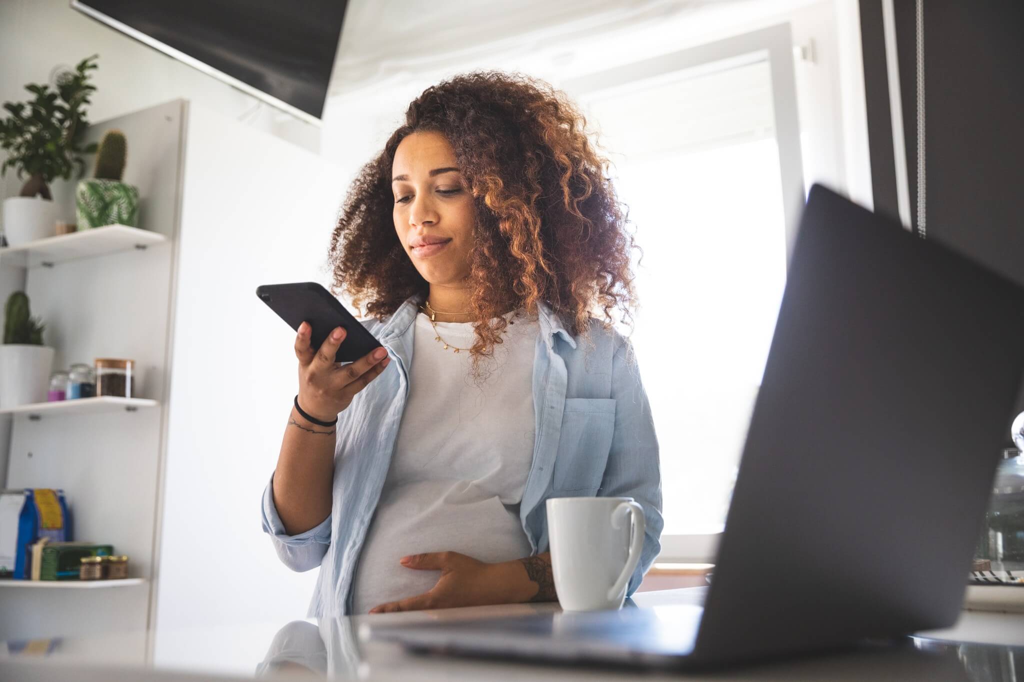 A woman with curly hair stands in a kitchen, looking at her phone.