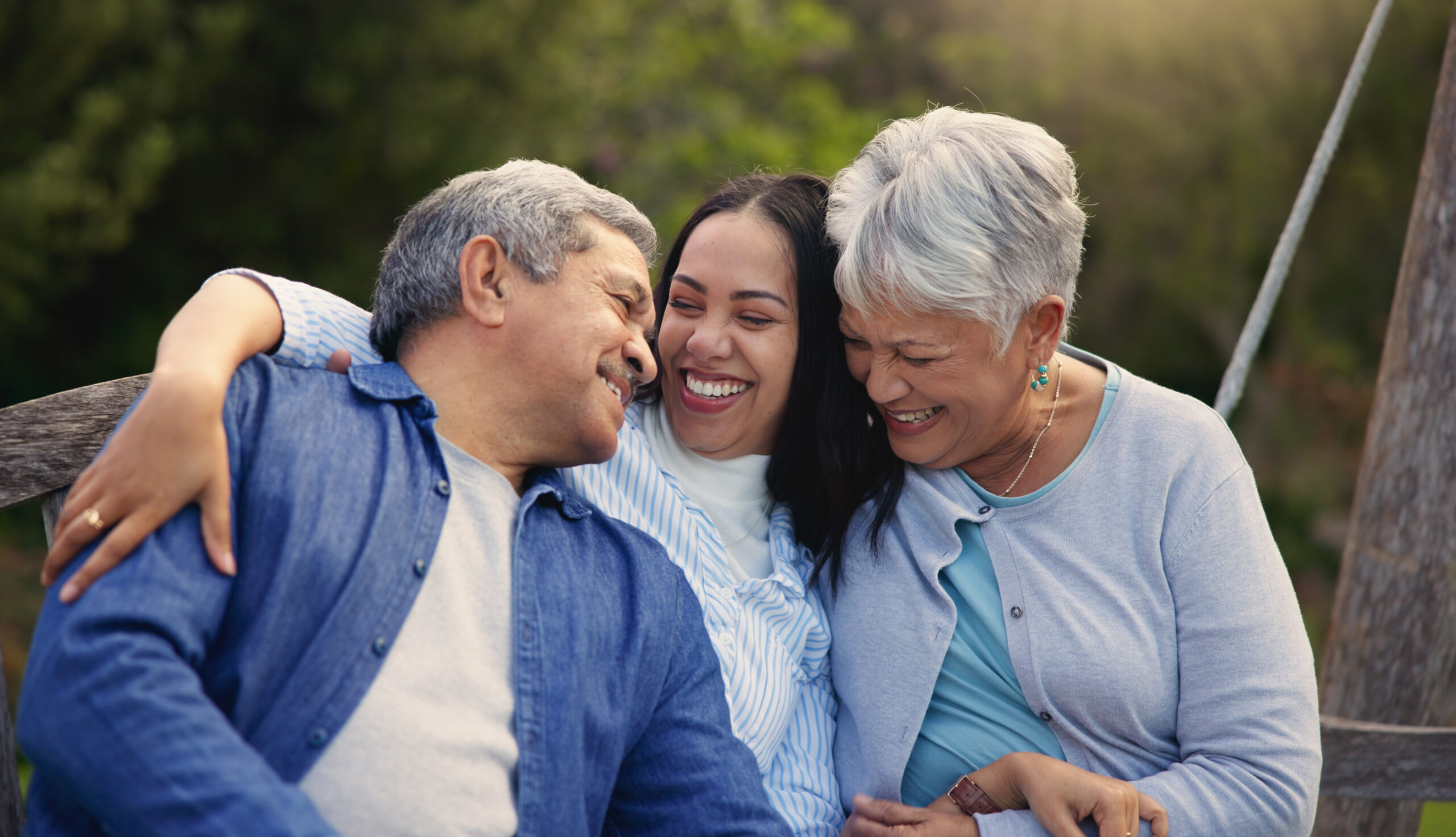 family of two seniors and younger woman sitting outside on a wooden bench hugging and smiling